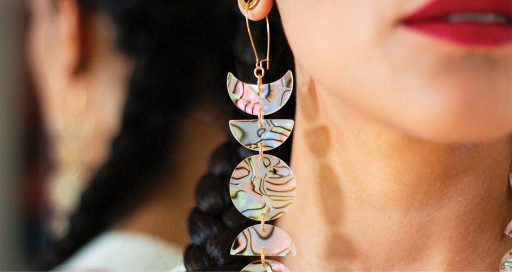 A laser cut earring hangs from the ear of an Indigenous femme.