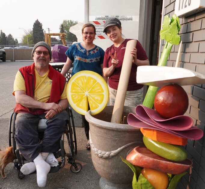 Dale Keith sits in his wheelchair next to two women standing. They are next to a sculpture of food.
