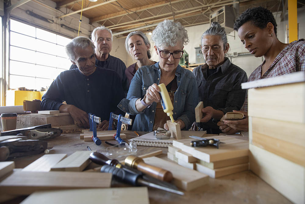 Five people look on as a woman glues two pieces of wood together in the Fuse33 woodshop.