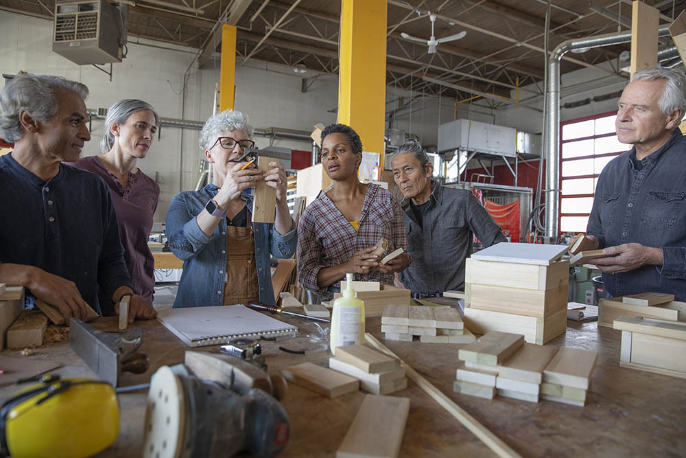 Five people look on as a woman glues two pieces of wood together in the Fuse33 woodshop.